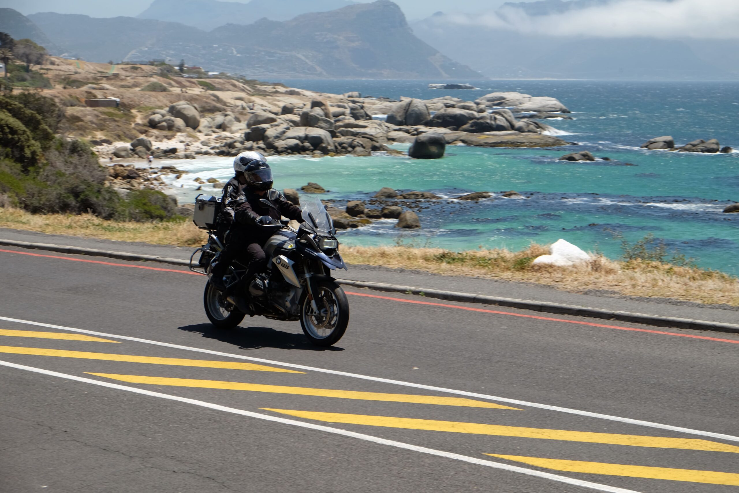 Motociclista e garupa em rodovia às margens de uma praia